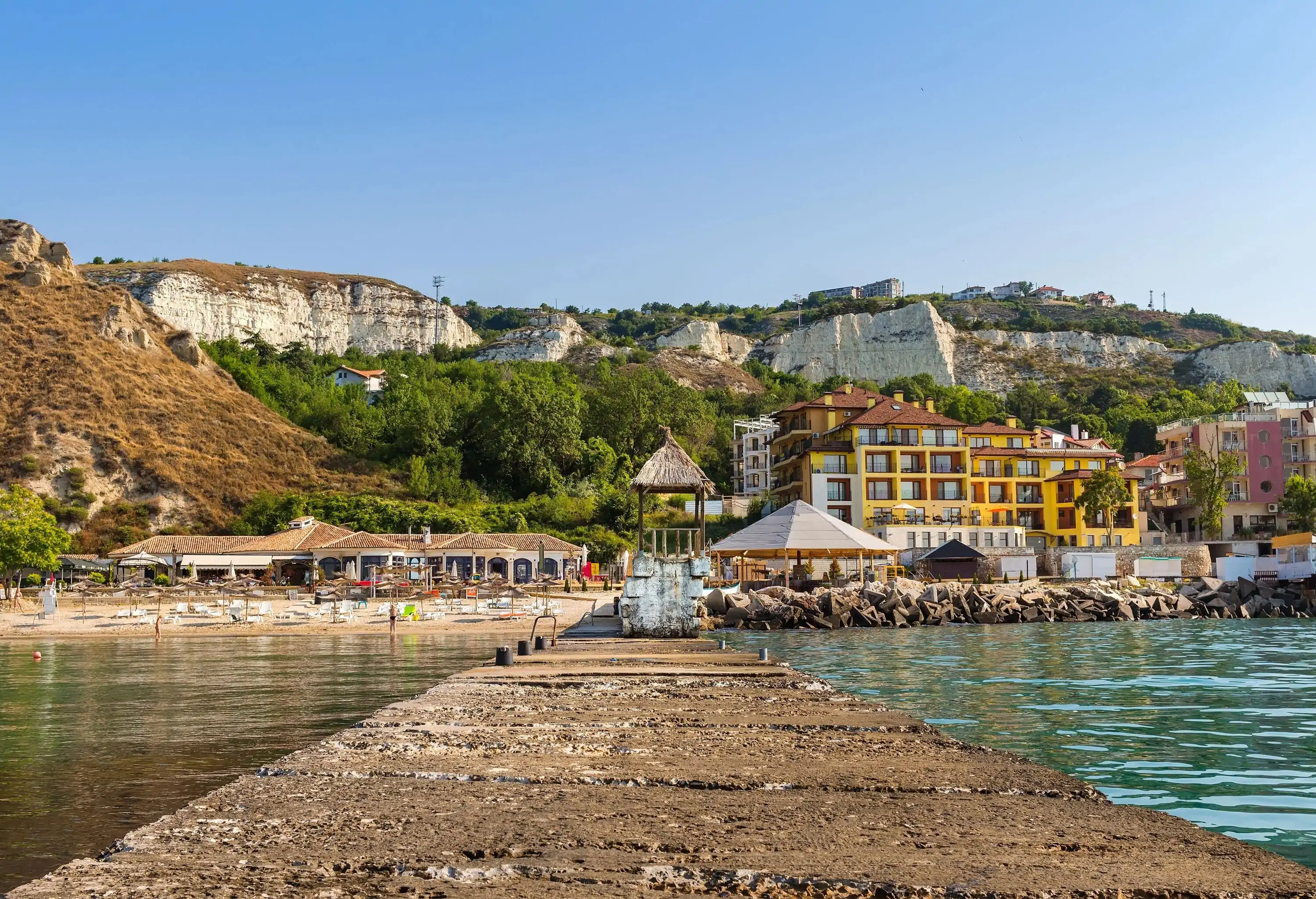 A coastal town and seaside resort with classic-looking buildings beneath the rocky hill overlooking the tranquil blue sea.