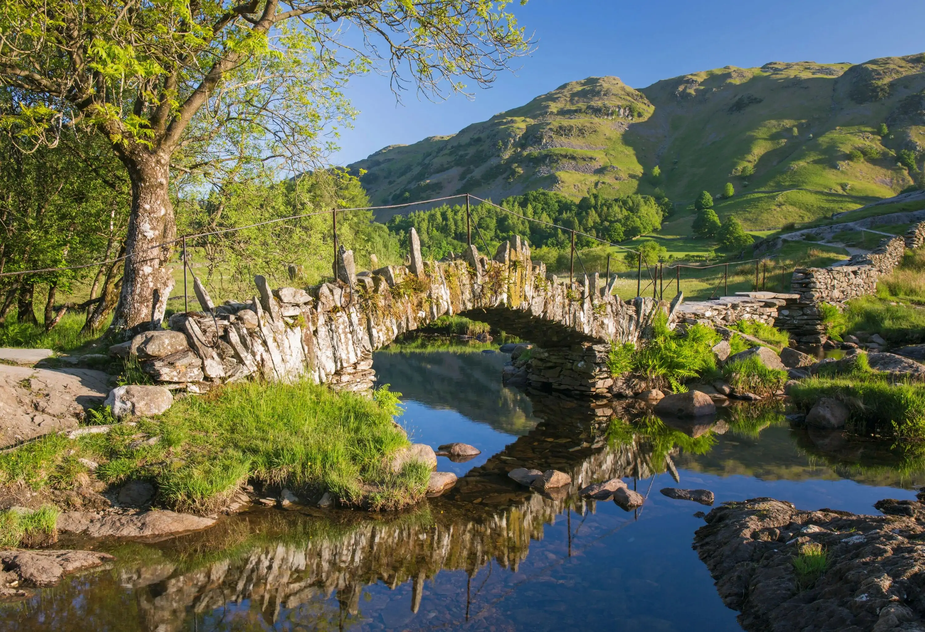 The surface of the river reflects a rock arch bridge and views of the surrounding mountains.