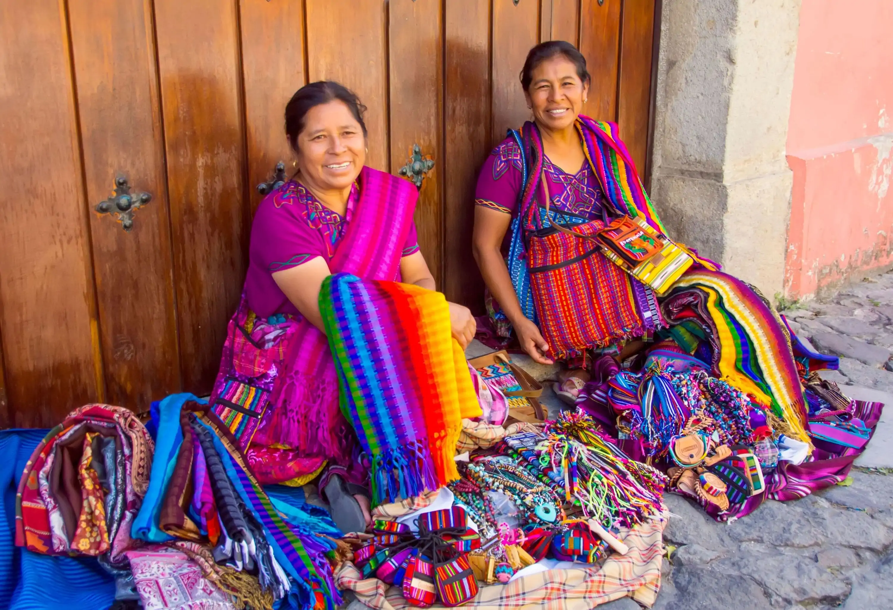 Two women wearing identical purple clothes selling textile handicrafts and souvenirs on a cobbled sidewalk.